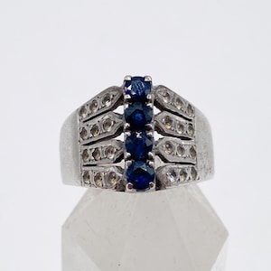 Puede incluir: Un anillo plateado con una fila vertical de cuatro piedras preciosas de color azul oscuro. La banda del anillo tiene un diseño rectangular con pequeñas piedras claras de acento. El anillo se muestra sobre una superficie clara y facetada.