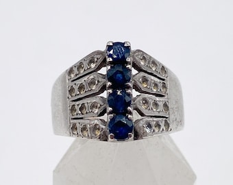 May include: A silver ring with a central line of four dark blue gemstones. The ring's band has a rectangular design with small, clear accent stones. The ring is displayed on a clear, faceted surface.