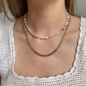May include: A gold chain necklace layered over a pearl necklace with gold accents. Both necklaces are on a model wearing a white crocheted top.