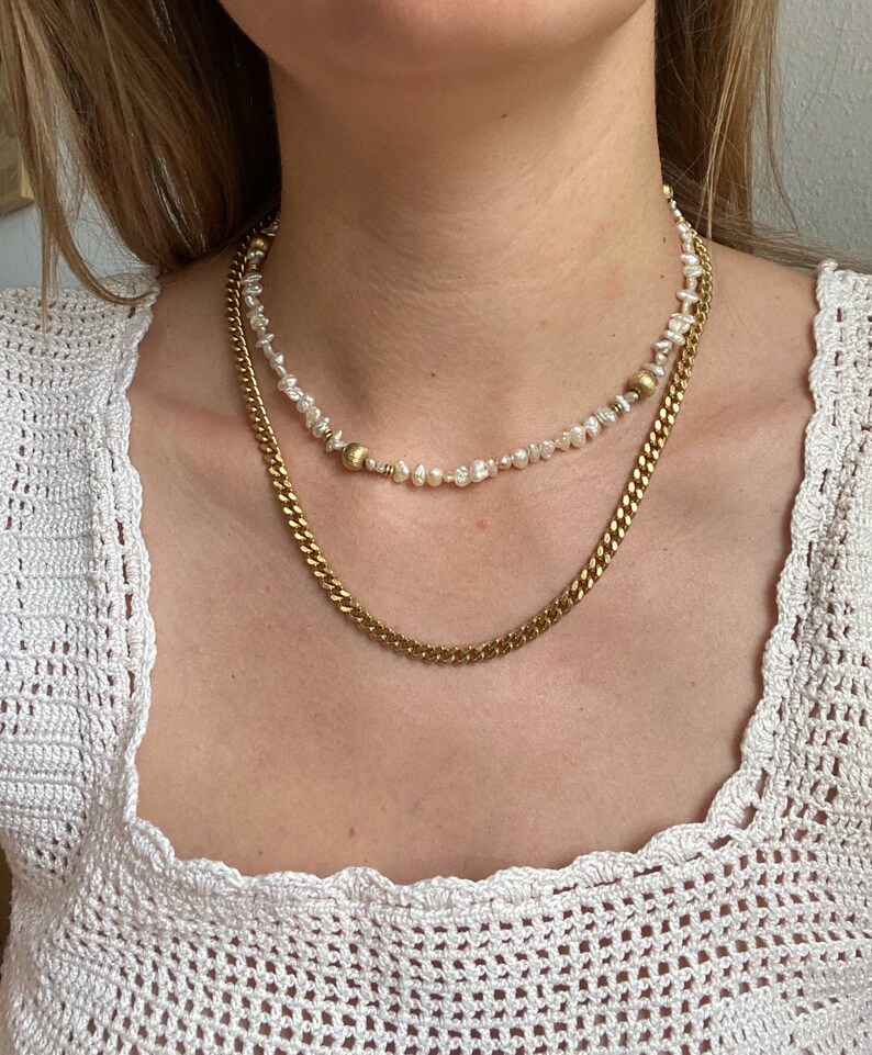 May include: A gold chain necklace layered over a pearl necklace with gold accents. Both necklaces are on a model wearing a white crocheted top.