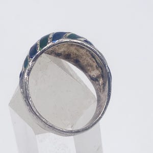 Puede incluir: Un anillo de plata con una banda que presenta secciones de esmalte azul y verde alternadas. El anillo se muestra desde un &aacute;ngulo lateral, destacando los colores y el dise&ntilde;o. El anillo est&aacute; colocado sobre una pantalla clara y geom&eacute;trica.