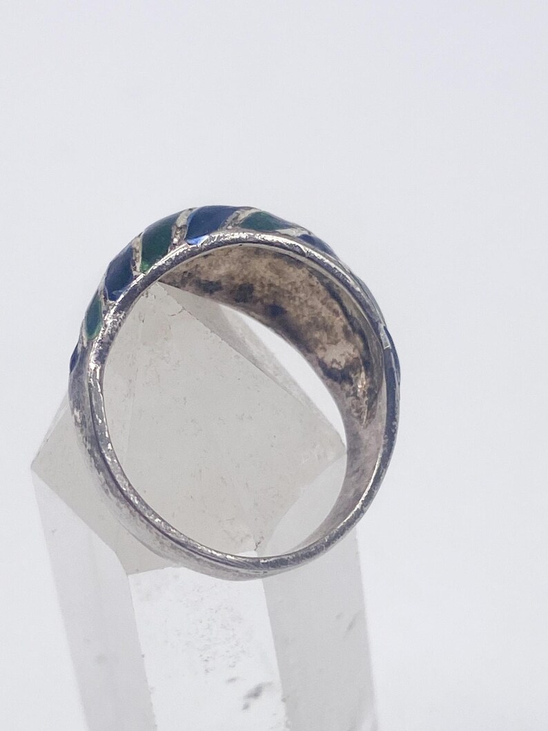 Puede incluir: Un anillo de plata con una banda que presenta secciones de esmalte azul y verde alternadas. El anillo se muestra desde un &aacute;ngulo lateral, destacando los colores y el dise&ntilde;o. El anillo est&aacute; colocado sobre una pantalla clara y geom&eacute;trica.