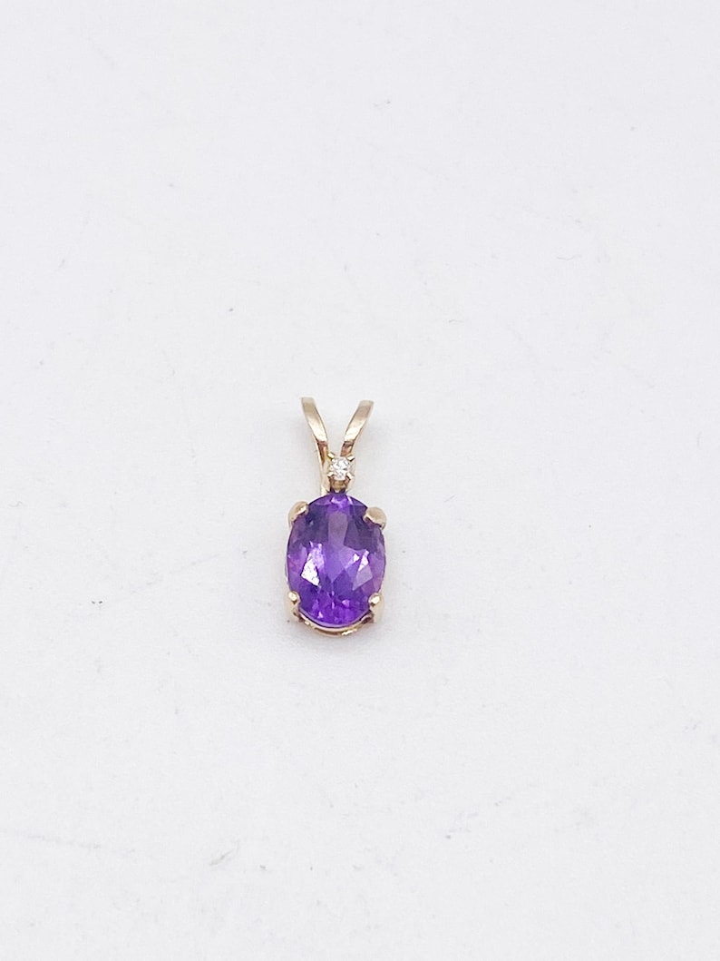 May include: A gold pendant featuring an oval, deep purple gemstone. The pendant has a small, round, clear stone set above the purple stone. The pendant is suspended from a gold bail.