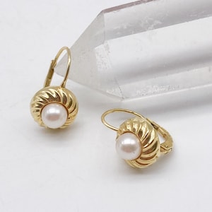 May include: A pair of gold-colored earrings with a pearl center. The earrings have a lever back closure and a textured, spiral design around the pearl. The earrings are displayed on a white surface.
