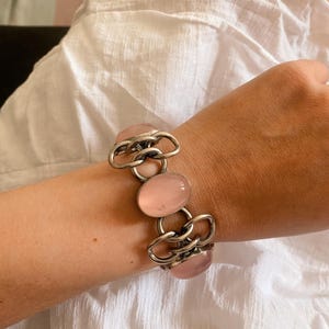 Puede incluir: Un brazalete plateado con tres piedras ovaladas de color rosa p&aacute;lido unidas por eslabones met&aacute;licos. El brazalete se muestra en una mu&ntilde;eca, destacando el dise&ntilde;o y el color de la joya.