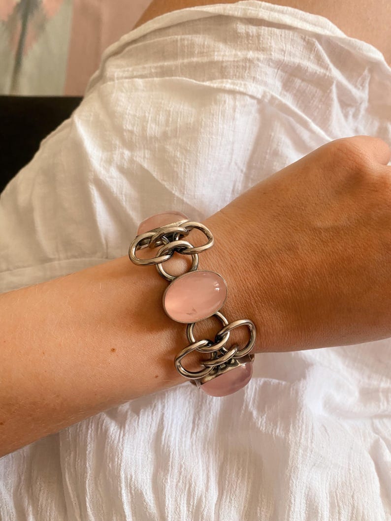 Puede incluir: Un brazalete plateado con tres piedras ovaladas de color rosa p&aacute;lido unidas por eslabones met&aacute;licos. El brazalete se muestra en una mu&ntilde;eca, destacando el dise&ntilde;o y el color de la joya.
