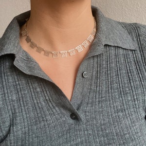 May include: A silver necklace with a geometric pattern of squares. The necklace is on a person wearing a gray shirt.