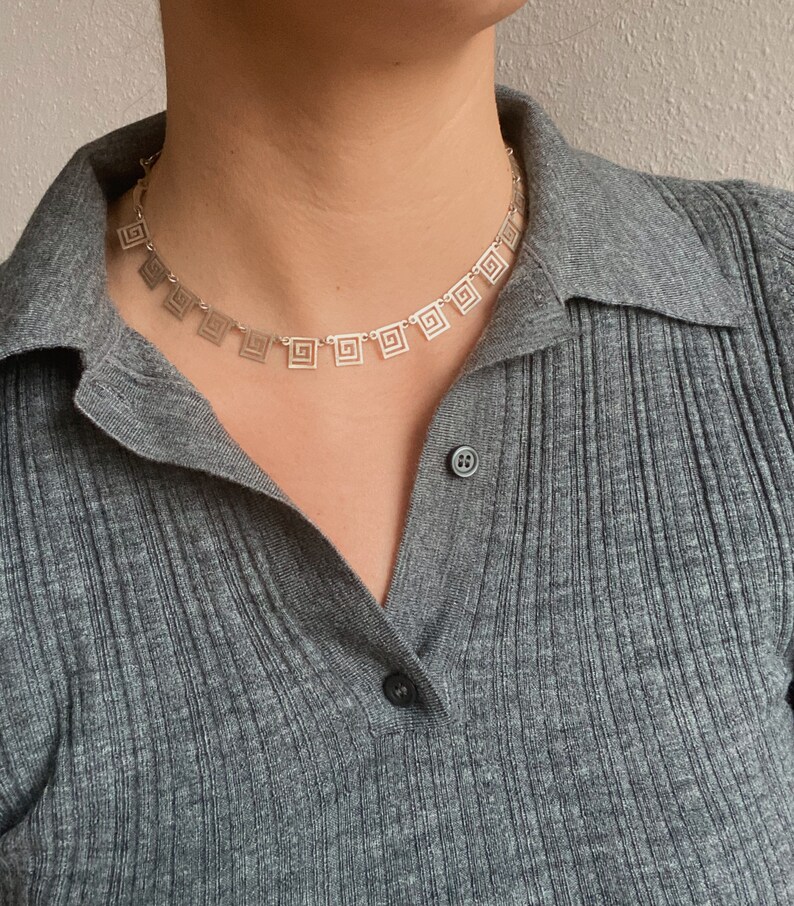 May include: A silver necklace with a geometric pattern of squares. The necklace is on a person wearing a gray shirt.