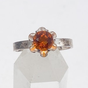 May include: A silver ring featuring a round, orange gemstone set in a flower-like design. The band is a simple, textured silver. The gemstone has a warm, amber hue, and the ring is displayed against a white background.