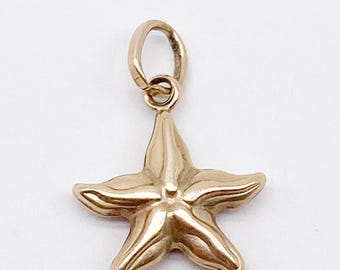 Puede incluir: Un dije de estrella de mar de color dorado, con cinco puntas y un pequeño lazo en la parte superior para sujetarlo a un collar o pulsera. El dije tiene una superficie lisa y pulida sobre un fondo blanco.