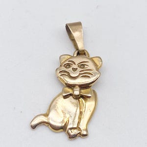 May include: A gold-colored cat pendant with a bow tie. The pendant features a smiling cat with detailed facial features and a loop at the top for a chain. The cat is sitting with its tail curled.
