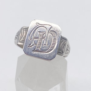 Puede incluir: Anillo de plata con una cara rectangular con un monograma grabado con las letras "AD". El anillo tiene una banda simple con diseños triangulares en los hombros. El anillo se muestra sobre una superficie cristalina transparente.
