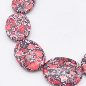 May include: A necklace composed of oval-shaped beads featuring a mosaic pattern of red, pink, and white stone fragments embedded in a dark gray matrix. The beads are strung together in a curved arrangement, showcasing a polished, smooth surface texture.