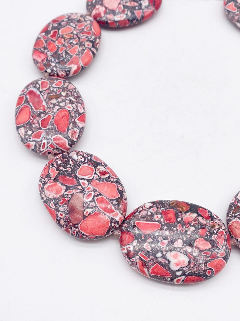 May include: A necklace composed of oval-shaped beads featuring a mosaic pattern of red, pink, and white stone fragments embedded in a dark gray matrix. The beads are strung together in a curved arrangement, showcasing a polished, smooth surface texture.