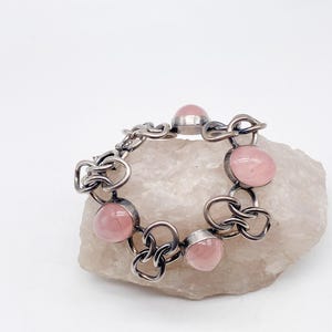 Puede incluir: Una pulsera plateada con seis gemas redondas de color rosa p&aacute;lido. La pulsera tiene un dise&ntilde;o de eslabones abiertos e entrelazados. Las gemas est&aacute;n engastadas en un bisel. La pulsera se exhibe sobre una superficie clara y texturizada.