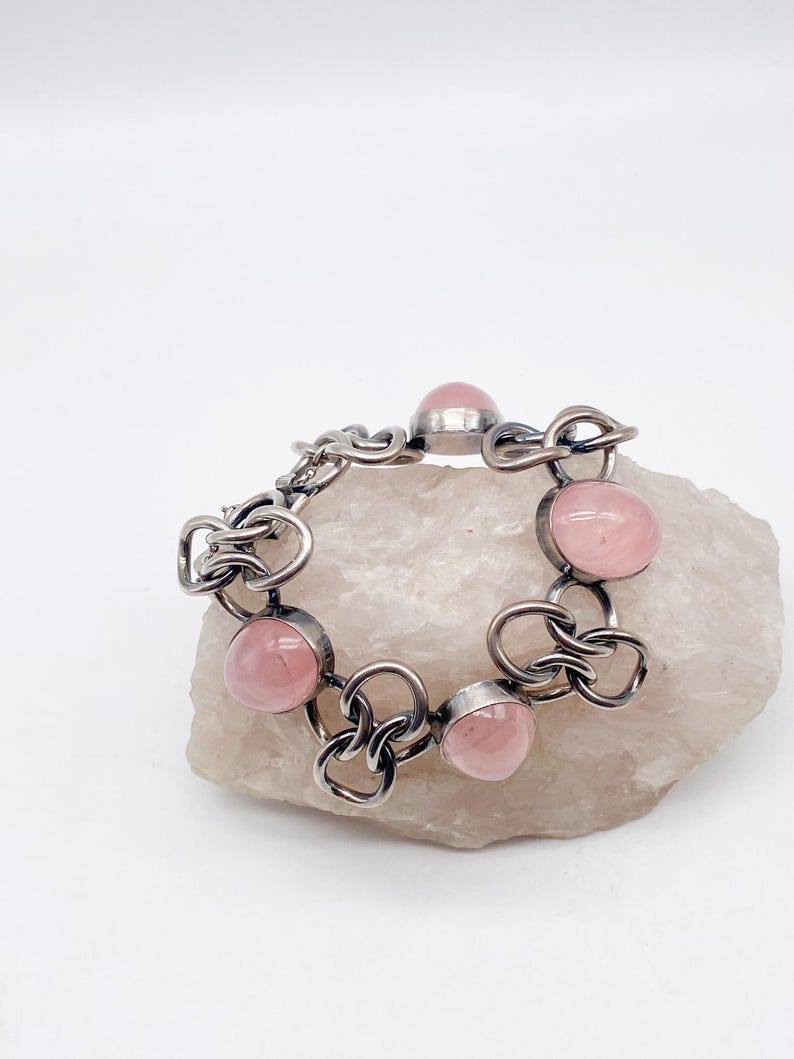 Puede incluir: Una pulsera plateada con seis gemas redondas de color rosa p&aacute;lido. La pulsera tiene un dise&ntilde;o de eslabones abiertos e entrelazados. Las gemas est&aacute;n engastadas en un bisel. La pulsera se exhibe sobre una superficie clara y texturizada.