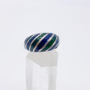 Puede incluir: Un anillo de plata con un dise&ntilde;o de esmalte a rayas azules y verdes. El anillo se exhibe sobre un soporte transparente y geom&eacute;trico sobre un fondo blanco. El dise&ntilde;o del anillo presenta bandas alternas de esmalte azul y verde.