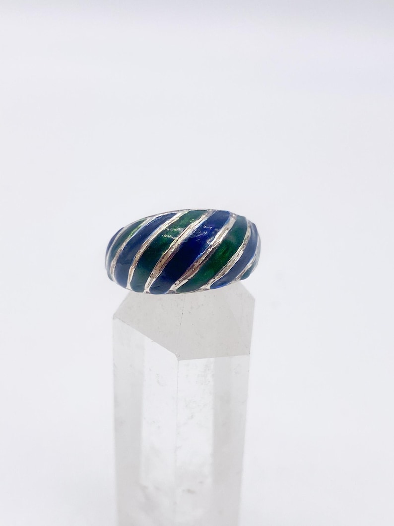 Puede incluir: Un anillo de plata con un dise&ntilde;o de esmalte a rayas azules y verdes. El anillo se exhibe sobre un soporte transparente y geom&eacute;trico sobre un fondo blanco. El dise&ntilde;o del anillo presenta bandas alternas de esmalte azul y verde.