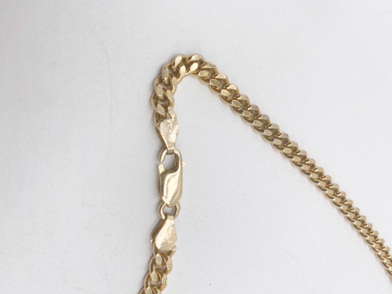 May include: A gold chain necklace with a curb link style. The chain is shown laying flat on a white surface.