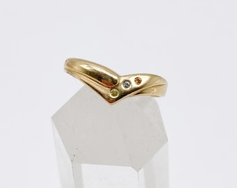 Op de afbeelding: Een goudkleurige ring met een V-vormig ontwerp. De ring heeft drie kleine edelstenen in geel, blauw en oranje. De ring wordt gepresenteerd op een heldere, facetgeslepen kristal. De ring is een sieraad.
