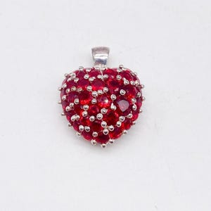 May include: A heart-shaped pendant featuring numerous round, faceted red stones set in a silver-toned metal frame. The stones are densely packed across the surface, held in place by small metal prongs, with a silver-toned bail at the top.