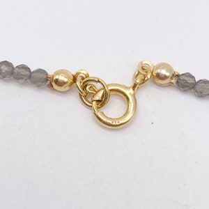 May include: A gold clasp with a round ring and a 925 stamp. The clasp is attached to a gray beaded necklace.