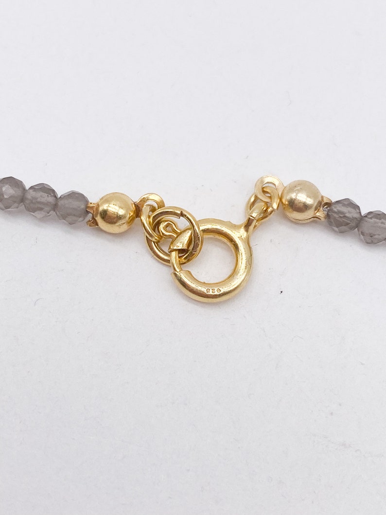 May include: A gold clasp with a round ring and a 925 stamp. The clasp is attached to a gray beaded necklace.