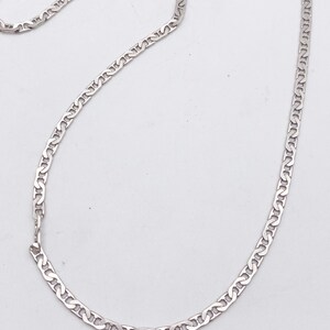 May include: A silver-colored chain necklace with a repeating, stylized link design. The necklace is laid out on a white surface, showcasing its delicate construction and reflective qualities. The chain is a classic jewelry piece.