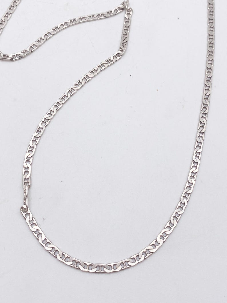 May include: A silver-colored chain necklace with a repeating, stylized link design. The necklace is laid out on a white surface, showcasing its delicate construction and reflective qualities. The chain is a classic jewelry piece.