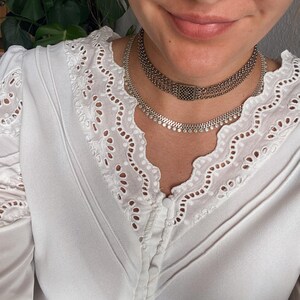 May include: A close-up of a person wearing two silver necklaces. The necklaces are layered, with one featuring a series of small, round embellishments and the other a chain design. The person is wearing a white blouse with eyelet lace detailing.