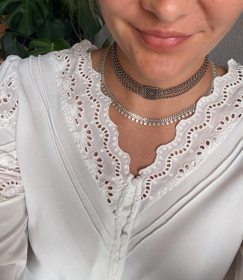 May include: A close-up of a person wearing two silver necklaces. The necklaces are layered, with one featuring a series of small, round embellishments and the other a chain design. The person is wearing a white blouse with eyelet lace detailing.