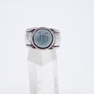 Può includere: Un anello d'argento con una pietra circolare azzurra. La pietra presenta un design stilizzato delle lettere "JJ" in blu più scuro. L'anello è posto su un supporto trasparente di cristallo su sfondo bianco.