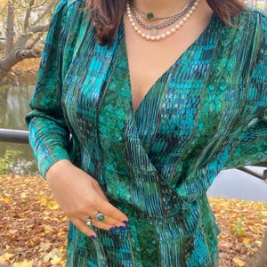 May include: A teal and green patterned wrap top and skirt set. The long-sleeved top has a V-neck and the skirt is visible at the bottom. A layered pearl necklace and rings with green stones are also visible.