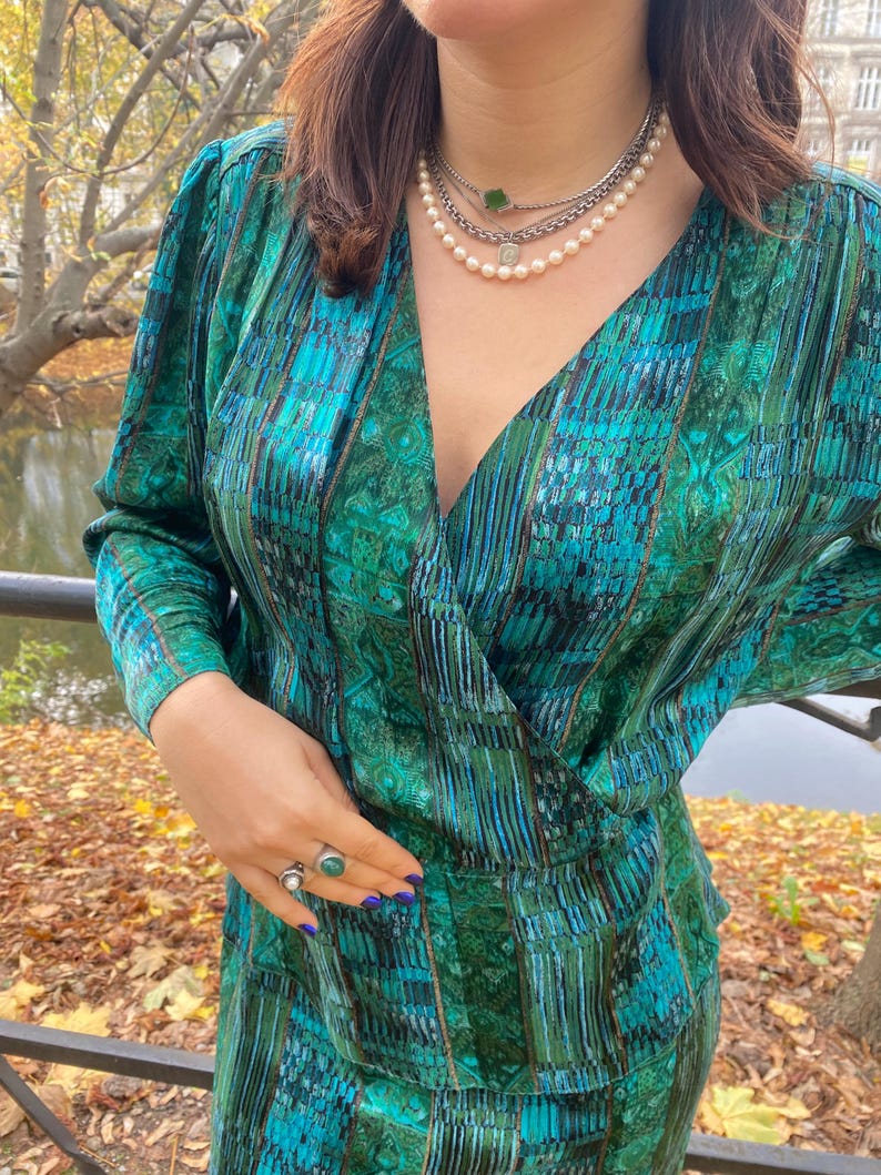 May include: A teal and green patterned wrap top and skirt set. The long-sleeved top has a V-neck and the skirt is visible at the bottom. A layered pearl necklace and rings with green stones are also visible.