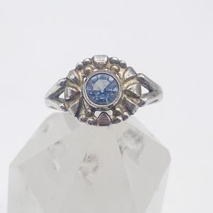 May include: A silver ring with a round, light blue gemstone at the center. The ring's setting features a floral design with small, raised details around the gemstone. The ring is displayed on a clear, faceted surface.