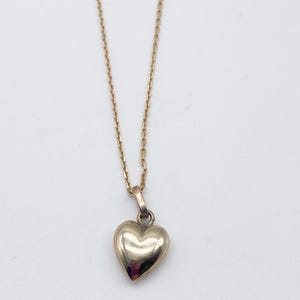 May include: A gold-toned necklace featuring a small, smooth, puffed heart-shaped pendant suspended from a delicate, thin metal chain. The pendant has a polished, reflective surface that catches the light, and the chain consists of small, uniform links.