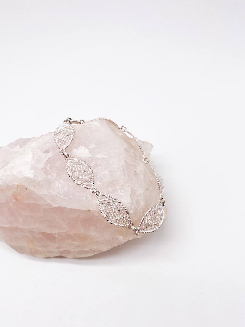 May include: A silver bracelet with a delicate, intricate design featuring a repeating pattern of oval-shaped links. The bracelet is displayed on a pink rock.