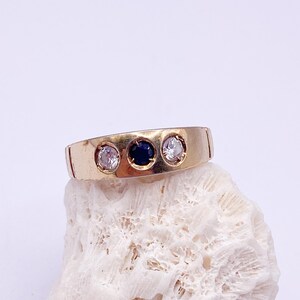 May include: A gold-toned ring with three stones: two clear and one dark blue. The ring is set on a white, textured surface, showcasing its design and the contrast of colors. The ring's band is a simple, flat design.