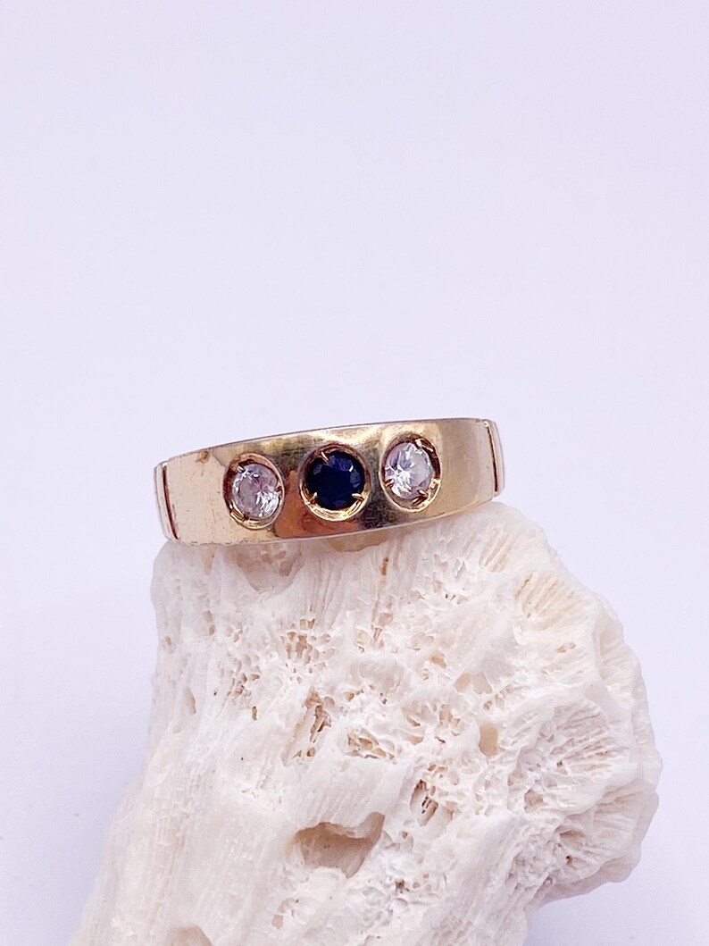 May include: A gold-toned ring with three stones: two clear and one dark blue. The ring is set on a white, textured surface, showcasing its design and the contrast of colors. The ring's band is a simple, flat design.