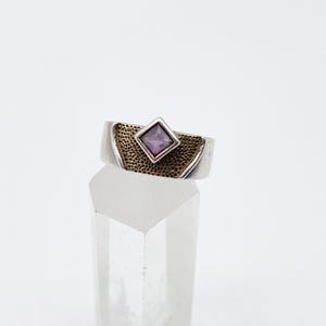 May include: A silver ring featuring a square, purple gemstone set within a textured, gold-toned band. The ring is displayed on a clear, crystal-like object against a white backdrop. The ring's design is simple yet elegant.