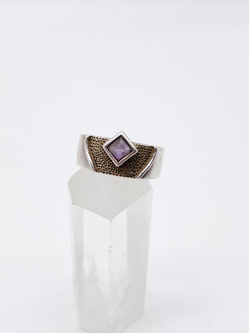 May include: A silver ring featuring a square, purple gemstone set within a textured, gold-toned band. The ring is displayed on a clear, crystal-like object against a white backdrop. The ring's design is simple yet elegant.