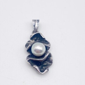 May include: A silver pendant featuring a large, round, white pearl nestled within a stylized, textured silver design. The pendant has a loop at the top for a chain. The design resembles a flower or abstract shape.