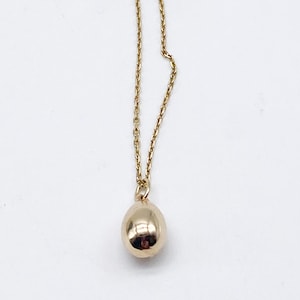 May include: A gold-colored necklace with a small, teardrop-shaped pendant. The pendant is smooth and reflective, hanging from a delicate chain. The necklace is set against a white background.
