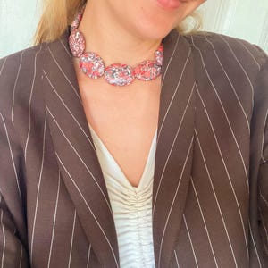 May include: A necklace featuring a series of oval-shaped beads with a mottled red, black, and white pattern. The necklace is worn over a white ruched top and a dark brown blazer with thin white pinstripes. The beads are arranged in a single strand.