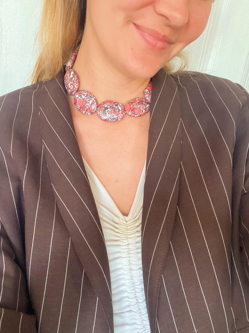 May include: A necklace featuring a series of oval-shaped beads with a mottled red, black, and white pattern. The necklace is worn over a white ruched top and a dark brown blazer with thin white pinstripes. The beads are arranged in a single strand.