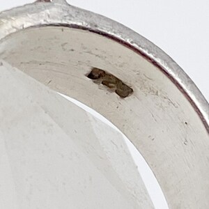 May include: Close-up of a silver ring with a smooth, polished surface. The ring has a slight curve and a small, raised detail on the top. The metal has a bright, reflective quality.