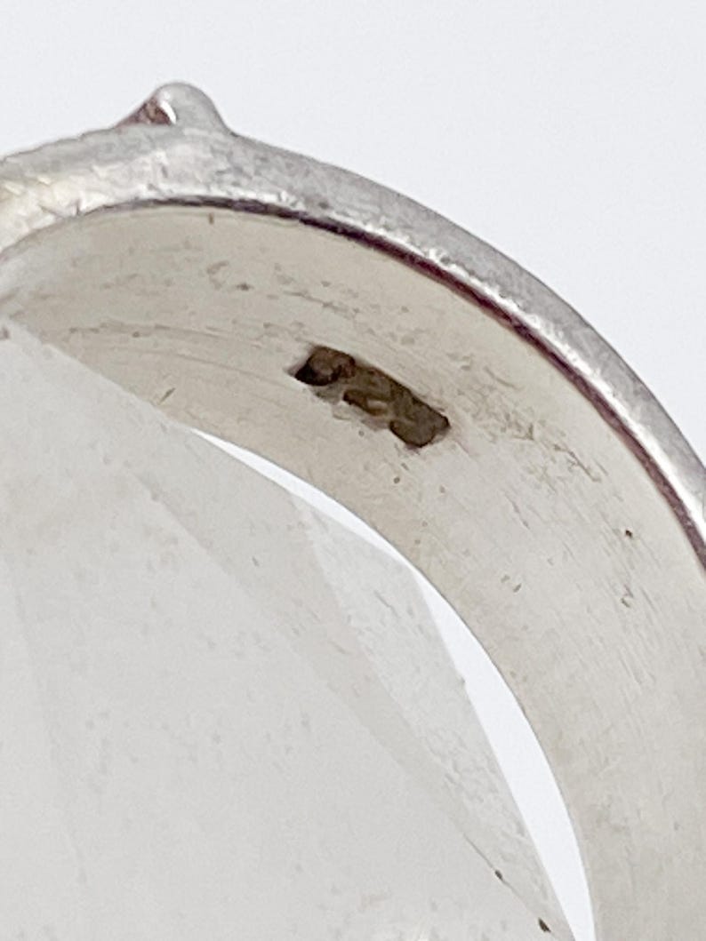May include: Close-up of a silver ring with a smooth, polished surface. The ring has a slight curve and a small, raised detail on the top. The metal has a bright, reflective quality.