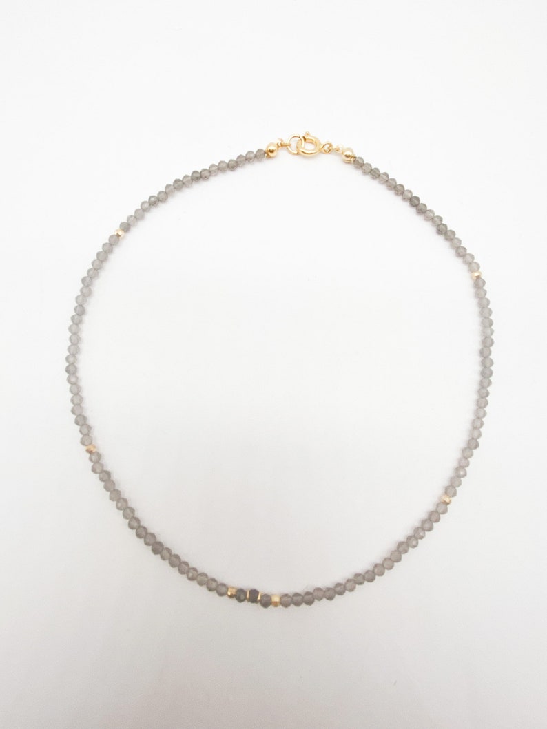 May include: A delicate gray beaded necklace with gold accents. The necklace is made of small, round beads and has a simple clasp.