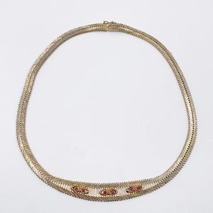 May include: A gold-coloured necklace with a herringbone chain pattern. The necklace has small, red gemstones set in a decorative design across the front. The clasp is visible at the back of the necklace.