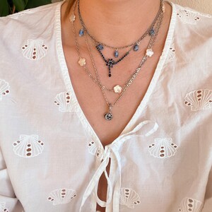 May include: A collection of layered necklaces in silver tones. The necklaces feature various pendants, including floral and gemstone designs. The model is wearing a white blouse with a seashell pattern.